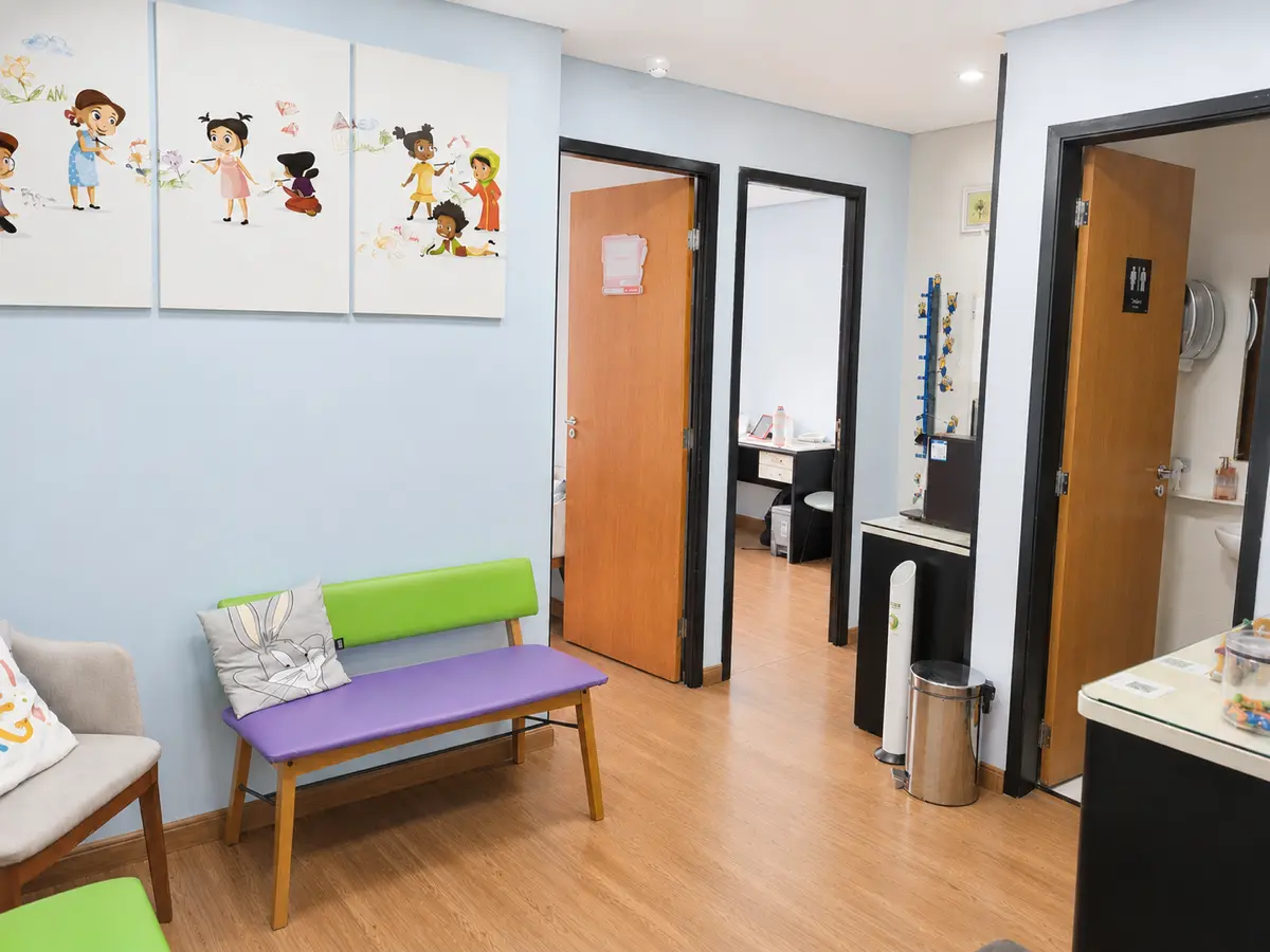 Sala de espera do consultório na Penha com paredes azuis, quadros infantis, banco roxo e verde com almofada do Pernalonga e piso de madeira